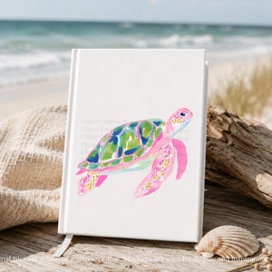 May include: A white notebook featuring a watercolour painting of a sea turtle. The turtle has pink flippers and head, and a shell with green, blue, and yellow patterns. The notebook is on a wooden surface near the beach.