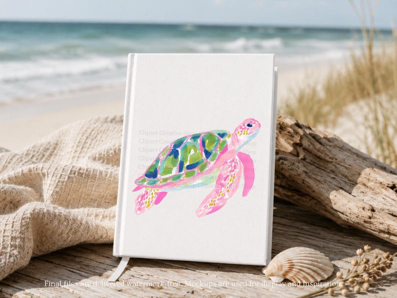 May include: A white notebook featuring a watercolour painting of a sea turtle. The turtle has pink flippers and head, and a shell with green, blue, and yellow patterns. The notebook is on a wooden surface near the beach.