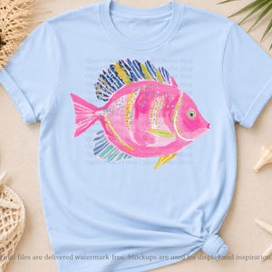 K&ouml;nnte beinhalten: Ein hellblaues Kurzarm-T-Shirt mit einer zentralen Grafik eines rosa Fisches mit gelb-blau gestreiften Flossen. Das Fischdesign enth&auml;lt kleine gelbe und blaue Sprenkel auf dem K&ouml;rper und ein einzelnes Auge auf der rechten Seite.