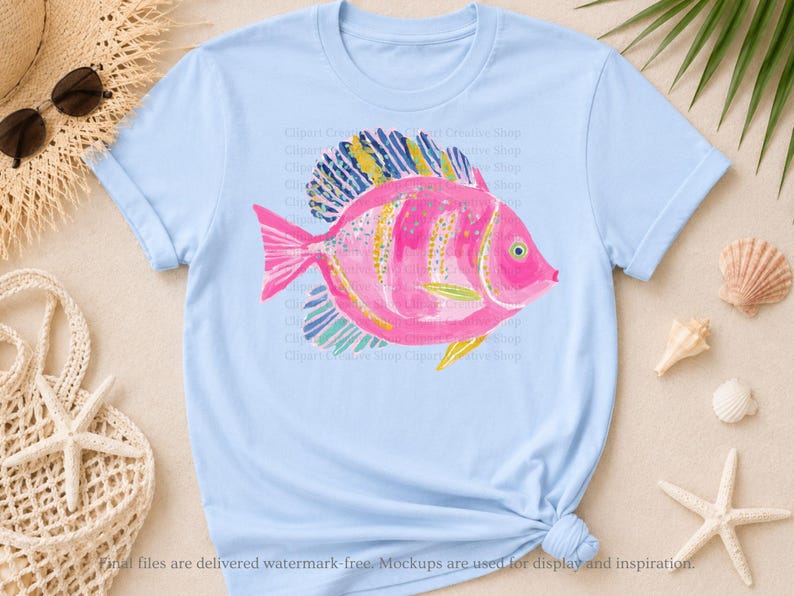 K&ouml;nnte beinhalten: Ein hellblaues Kurzarm-T-Shirt mit einer zentralen Grafik eines rosa Fisches mit gelb-blau gestreiften Flossen. Das Fischdesign enth&auml;lt kleine gelbe und blaue Sprenkel auf dem K&ouml;rper und ein einzelnes Auge auf der rechten Seite.
