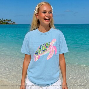 May include: A light blue t-shirt with a watercolour sea turtle design. The turtle has a green and blue shell and pink flippers. The shirt is worn by a person on a beach with turquoise water and a blue sky.