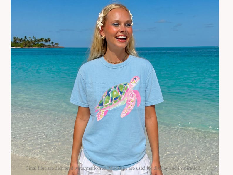 May include: A light blue t-shirt with a watercolour sea turtle design. The turtle has a green and blue shell and pink flippers. The shirt is worn by a person on a beach with turquoise water and a blue sky.