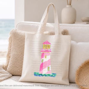 K&ouml;nnte beinhalten: Eine cremefarbene Tragetasche mit einer Aquarellillustration eines rosa und wei&szlig; gestreiften Leuchtturms mit gelben Akzenten. Der Leuchtturm steht auf einem blauen Sockel mit einem rosa Dach. Die Tasche liegt auf einem wei&szlig;en Sofa.
