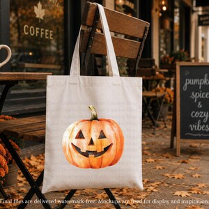 May include: A cream-colored canvas tote bag featuring a centered, vibrant orange jack-o'-lantern illustration with a carved triangular nose, eyes, and a wide, jagged grin. The pumpkin has a green stem and is rendered in a watercolor-style aesthetic.