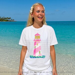 K&ouml;nnte beinhalten: Wei&szlig;es T-Shirt mit einer Aquarellillustration eines rosa und wei&szlig; gestreiften Leuchtturms mit gelbem Dach. Das T-Shirt wird von einer Person an einem Strand mit t&uuml;rkisfarbenem Wasser und blauem Himmel getragen. Der Text "Final files are delivered watermark-free. Mockups are used for display and inspiration."