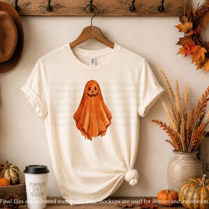 Puede incluir: Una camiseta de manga corta de color crema con una ilustraci&oacute;n central estilo acuarela de un fantasma naranja con una cara de calabaza tallada. La camiseta tiene cuello redondo y se muestra en una percha de madera sobre una pared.