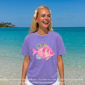 K&ouml;nnte beinhalten: Ein lavendelfarbenes Kurzarm-T-Shirt mit einem Grafikdruck eines rosa Fisches mit gelben und blauen Flossendetails. Die Fischillustration ist mittig auf der Brust platziert. Das Kleidungsst&uuml;ck hat einen Rundhalsausschnitt und eine Standardpassform.