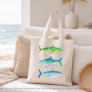 May include: A beige tote bag featuring three colorful watercolor fish illustrations. The fish are green, blue, and blue with pink accents. The bag is on a white couch with pillows and a straw hat.