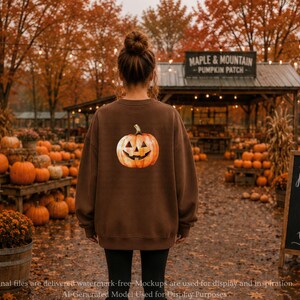 May include: A brown crewneck sweatshirt featuring a graphic of a carved jack-o'-lantern pumpkin on the back. The sweatshirt is worn over black leggings, with the pumpkin design centered on the upper back area of the garment.