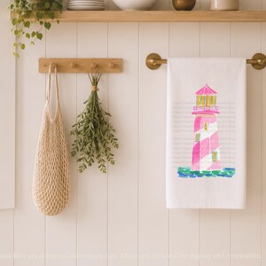 K&ouml;nnte beinhalten: Wei&szlig;es K&uuml;chentuch mit einem rosa und wei&szlig;en Leuchtturm-Design. Der Leuchtturm hat ein rosa Dach und ist vor einem blau-gr&uuml;nen Aquarellmeer platziert. Das Handtuch h&auml;ngt an einer Messingstange.