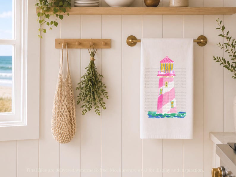 K&ouml;nnte beinhalten: Wei&szlig;es K&uuml;chentuch mit einem rosa und wei&szlig;en Leuchtturm-Design. Der Leuchtturm hat ein rosa Dach und ist vor einem blau-gr&uuml;nen Aquarellmeer platziert. Das Handtuch h&auml;ngt an einer Messingstange.
