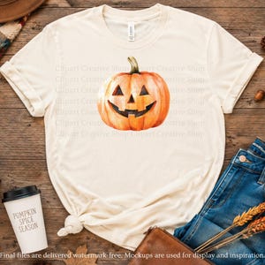 May include: A cream-colored short-sleeve t-shirt featuring a central graphic of an orange jack-o'-lantern pumpkin with a carved smiling face. The shirt has a crew neckline and is displayed flat, showcasing the pumpkin illustration clearly.
