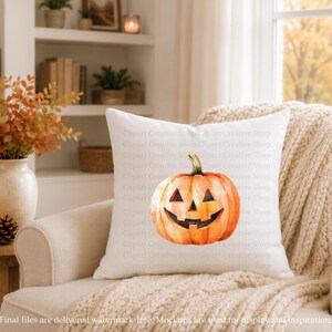 May include: A square white throw pillow featuring a centered watercolor-style illustration of an orange jack-o'-lantern pumpkin with a carved triangular nose, eyes, and a wide, jagged grin, set against a plain, light-colored fabric background.