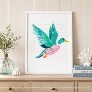 Op de afbeelding: Een ingelijste kunstdruk met een aquarelillustratie van een vliegende eend met levendige blauwgroene vleugels, een roze lichaam met gouden accenten en een gele snavel. Het kunstwerk staat in een witte lijst op een houten ondergrond.
