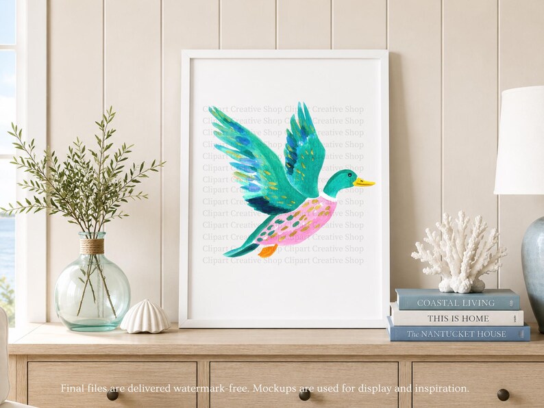 Op de afbeelding: Een ingelijste kunstdruk met een aquarelillustratie van een vliegende eend met levendige blauwgroene vleugels, een roze lichaam met gouden accenten en een gele snavel. Het kunstwerk staat in een witte lijst op een houten ondergrond.