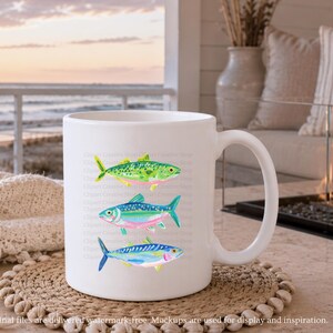 May include: White ceramic mug featuring three colorful watercolor fish illustrations. The fish are in shades of green, blue, pink, and yellow. The mug is on a woven coaster, with a beach scene in the background. The mug has a curved handle.