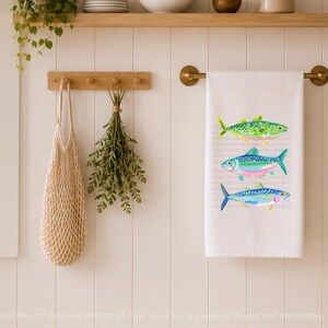 May include: White kitchen towel with three colorful fish illustrations. The fish are green, blue, and yellow, with pink and blue accents. The towel hangs on a brass rod. A wooden peg rack holds a net bag and greenery.