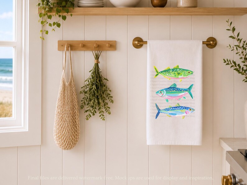 May include: White kitchen towel with three colorful fish illustrations. The fish are green, blue, and yellow, with pink and blue accents. The towel hangs on a brass rod. A wooden peg rack holds a net bag and greenery.