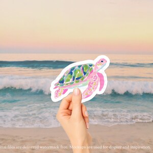May include: A watercolour sticker of a sea turtle with a pink body and green and blue shell. The sticker is held in front of a beach scene with the ocean and a sunset in the background.