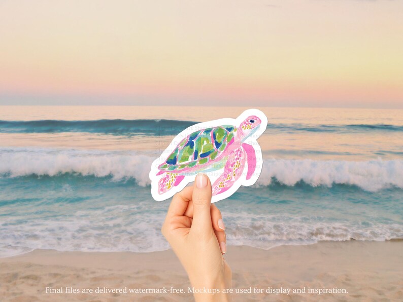 May include: A watercolour sticker of a sea turtle with a pink body and green and blue shell. The sticker is held in front of a beach scene with the ocean and a sunset in the background.