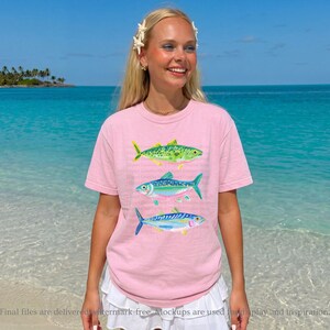 May include: A light pink t-shirt featuring three colorful fish illustrations. The fish are green, blue, and blue with yellow accents. The shirt is worn by a person standing on a beach with a blue ocean in the background.