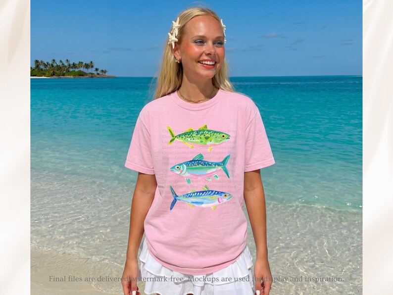 May include: A light pink t-shirt featuring three colorful fish illustrations. The fish are green, blue, and blue with yellow accents. The shirt is worn by a person standing on a beach with a blue ocean in the background.
