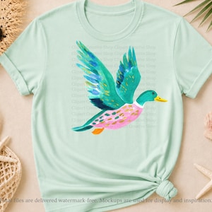 Op de afbeelding: Een licht mintgroen T-shirt met korte mouwen en een centrale afbeelding van een vliegende eend. De eend is afgebeeld met levendige blauwgroene vleugels, een roze lichaam met groene stippen en een gele snavel in een aquarelachtige stijl.