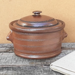 以下が含まれることがあります： 蓋付きの丸い茶色の陶器製キャセロール皿。本体と蓋の周囲には、ロープのような水平のエンボス加工が施されています。鍋には2つの小さなサイドハンドルと、蓋の中央にノブが付いており、滑らかな釉薬仕上げが特徴です。