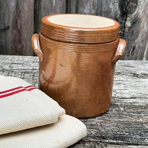 Può includere: Un vaso in ceramica smaltata marrone con due manici laterali e un coperchio abbinato, posizionato accanto a un canovaccio da cucina color crema piegato, caratterizzato da due strisce rosse parallele che corrono orizzontalmente sul tessuto.
