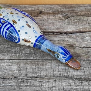 Op de afbeelding: Een handbeschilderd keramieken eendenbeeldje met een wit lichaam en blauwe en groene patronen, waaronder stippen en bladvormen. De eend heeft een blauwe kop met gedetailleerde markeringen rond de ogen en een bruine snavel met een glanzende afwerking.