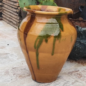 Può includere: Un vaso in ceramica smaltata con un bordo largo e svasato e una base affusolata. L'esterno presenta un colore di base beige caldo con evidenti colature verticali di smalto marrone scuro e verde intenso che scendono lungo le pareti.