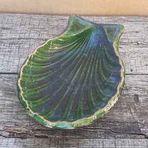 May include: A shell-shaped ceramic dish featuring a deep green glaze with dark blue accents. The bowl has a scalloped edge with a visible light-colored rim, showcasing a textured, ribbed surface that mimics the natural form of a seashell.