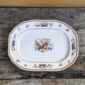 Op de afbeelding: Een ovale witte keramische serveerschaal met een centraal kleurrijk fruitmotief van appels, druiven en pruimen. De rand is versierd met een herhalend patroon van kleine fruitgroepen, vlinders en een goudkleurige gevlochten rand.