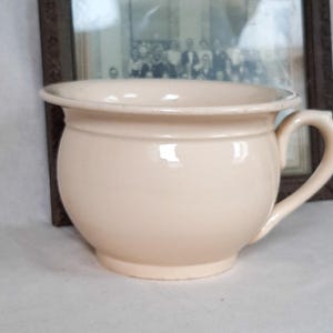 Op de afbeelding: Een crèmekleurige keramische kamerpot met een brede, ronde kom, een uitlopende rand en een enkel gebogen handvat aan de zijkant. Het oppervlak van het vat is glad en glanzend en heeft een uniforme lichtbeige tint over de gehele buitenkant.