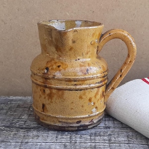 Könnte beinhalten: Ein senfgelber Keramikkrug mit einer glänzenden, gesprenkelten Glasur und einem einzelnen geschwungenen Henkel. Der Krug hat einen breiten, zylindrischen Körper mit zwei horizontalen Rillen um die Basis und einen kleinen Ausguss am oberen Rand.