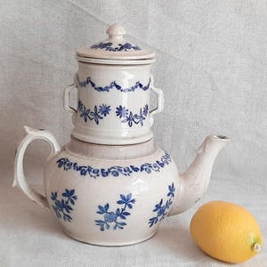 Puede incluir: Una tetera de cerámica blanca con tapa apilada y patrones florales azules. La tetera presenta un cuerpo redondeado, un asa curva, un pico vertedor y una tapa a juego con un pomo. Un limón amarillo está colocado junto a la tetera.