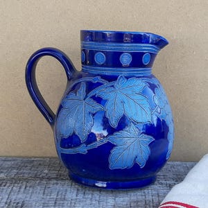 May include: A deep cobalt blue ceramic pitcher featuring a prominent handle and a wide spout. The body of the pitcher is decorated with embossed light blue maple leaf patterns and a band of circular motifs around the upper neck area.