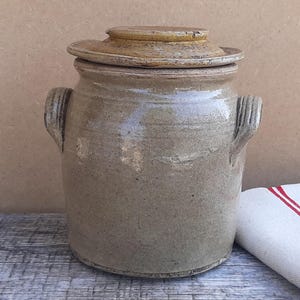 May include: A cylindrical stoneware crock with a glossy, light beige glaze and a matching lid. The vessel features two vertical, ribbed handles on opposite sides. The surface shows subtle variations in colour and texture, characteristic of glazed pottery.
