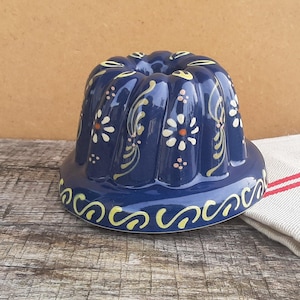 May include: A dark blue ceramic bundt cake mold featuring a fluted design and hand-painted floral patterns in cream and orange. The base of the mold is decorated with a continuous yellow scroll motif, and the surface has a glossy finish.