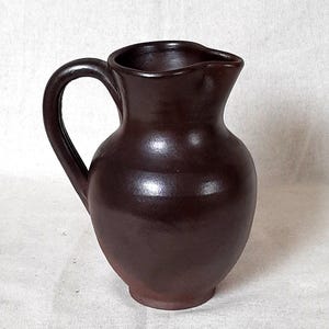Puede incluir: Una jarra de cerámica marrón oscuro con un asa curva y un pico vertedor. La jarra tiene un cuerpo redondeado y una base ligeramente ensanchada. La superficie tiene un acabado brillante. La jarra se utiliza probablemente para servir bebidas.