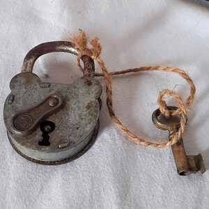 May include: An antique, weathered gray padlock with a key attached by a natural fiber cord. The lock has a rounded shape and a keyhole. The key is a tarnished brass color. A portion of a gold-colored clock is visible in the background.