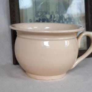 May include: A cream-colored ceramic chamber pot with a rounded body, a wide flared rim, and a single curved handle on the side. The vessel has a smooth, glossy finish and a simple, functional design with a circular base and a deep interior basin.