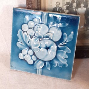 Könnte beinhalten: Quadratische, blaue Keramikfliese mit einem erhabenen Reliefdesign aus Früchten und Blättern. Die Fliese hat eine glänzende Oberfläche und ist wahrscheinlich Vintage. Die Früchte scheinen Trauben und Äpfel zu sein. Die Fliese ist vor einem neutralen Hintergrund platziert.
