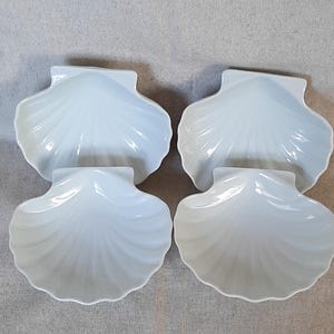 Könnte beinhalten: Ein Set aus vier weißen Keramikschalen in Form von Jakobsmuscheln. Jede Schale hat eine glänzende Oberfläche mit geprägten radialen Rillen, die vom Scharnierbereich zum gewellten Rand verlaufen und ein einheitliches, symmetrisches Aussehen erzeugen.