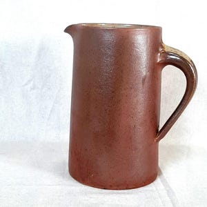 Peut inclure: Une cruche en céramique marron avec une anse et un bec verseur. La cruche a une finition lisse et mate et une forme légèrement conique. La poignée est d'une nuance de brun plus foncée. Elle est probablement utilisée pour verser des liquides.