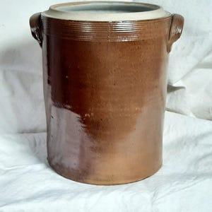 May include: A brown stoneware crock with a cylindrical shape and two side handles. The crock has a light-colored rim and a glossy finish. This antique kitchenware item is suitable for storage or decoration.