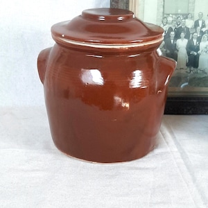 Può includere: Un vaso di ceramica marrone con coperchio, con due manici laterali. Il vaso ha una finitura lucida e un bordo bianco sul coperchio. Sullo sfondo c'è una fotografia in bianco e nero incorniciata.
