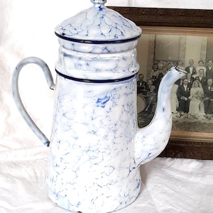 May include: A vintage enamelware coffee pot in white with a blue marbled pattern. The pot has a lid and handle, with dark blue trim. The pot is on a white surface. The pot is likely from the mid-20th century.