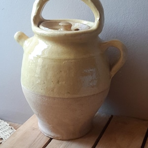 Può includere: Una brocca in ceramica smaltata gialla con un grande manico ad arco sulla parte superiore, una piccola impugnatura laterale e un coperchio abbinato. La parte superiore presenta una finitura gialla lucida, mentre la base è in argilla opaca.