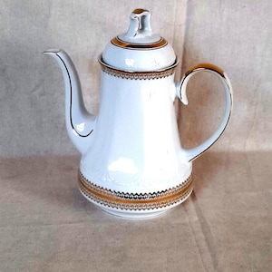 Puede incluir: Cafetera de porcelana blanca con boquilla y asa curvadas. La cafetera presenta ribetes dorados en el borde, la base y el asa, con una banda dorada decorativa en el centro. La tapa también es blanca con detalles dorados.